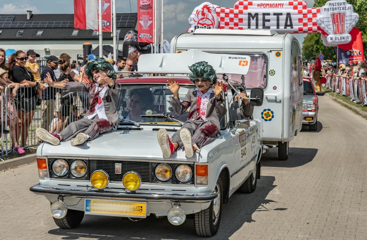 Fiat 125p z przyczepą kempingową na mecie Rajdu Koguta w Oławie — dwóch przebierańców w zielonych perukach siedzi na masce, tłum kibiców za barierkami