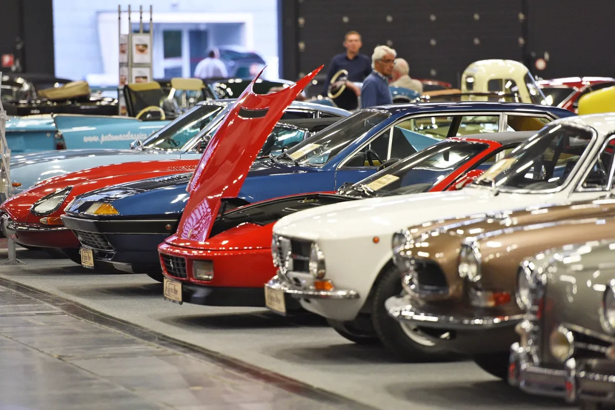 Rząd klasycznych samochodów na targach w hali wystawowej — widoczne m.in. Fiat Dino Coupé, Volvo P1800 i inne europejskie klasyki lat sześćdziesiątych i siedemdziesiątych