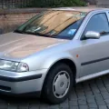 Srebrny liftback przy ceglanym murze — pojedyncze reflektory i chromowana atrapa zdradzają wersję sprzed liftingu 2000 roku, najprostszą twarz pierwszej Octavii. Samochód bez pretensji, budowany po to, żeby jeździć, a nie robić wrażenie.
