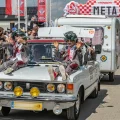 Fiat 125p z przyczepą kempingową na mecie Rajdu Koguta w Oławie — dwóch przebierańców w zielonych perukach siedzi na masce, tłum kibiców za barierkami