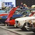 Rząd klasycznych samochodów na targach w hali wystawowej — widoczne m.in. Fiat Dino Coupé, Volvo P1800 i inne europejskie klasyki lat sześćdziesiątych i siedemdziesiątych