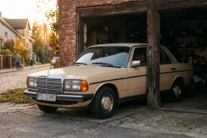 Beżowy Mercedes W123 w progu garażu — pół w słońcu, pół w cieniu. Decyzja o żółtych tablicach wygląda dokładnie tak.