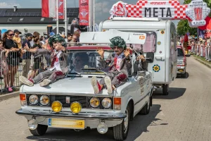 Fiat 125p z przyczepą kempingową na mecie Rajdu Koguta w Oławie — dwóch przebierańców w zielonych perukach siedzi na masce, tłum kibiców za barierkami