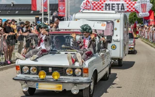 Fiat 125p z przyczepą kempingową na mecie Rajdu Koguta w Oławie — dwóch przebierańców w zielonych perukach siedzi na masce, tłum kibiców za barierkami