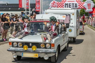 Fiat 125p z przyczepą kempingową na mecie Rajdu Koguta w Oławie — dwóch przebierańców w zielonych perukach siedzi na masce, tłum kibiców za barierkami