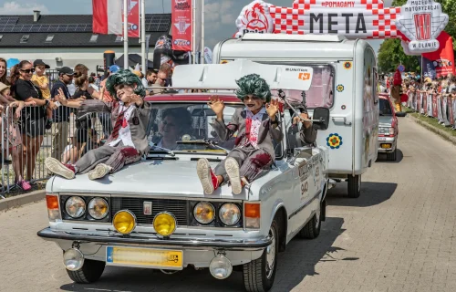 Fiat 125p z przyczepą kempingową na mecie Rajdu Koguta w Oławie — dwóch przebierańców w zielonych perukach siedzi na masce, tłum kibiców za barierkami