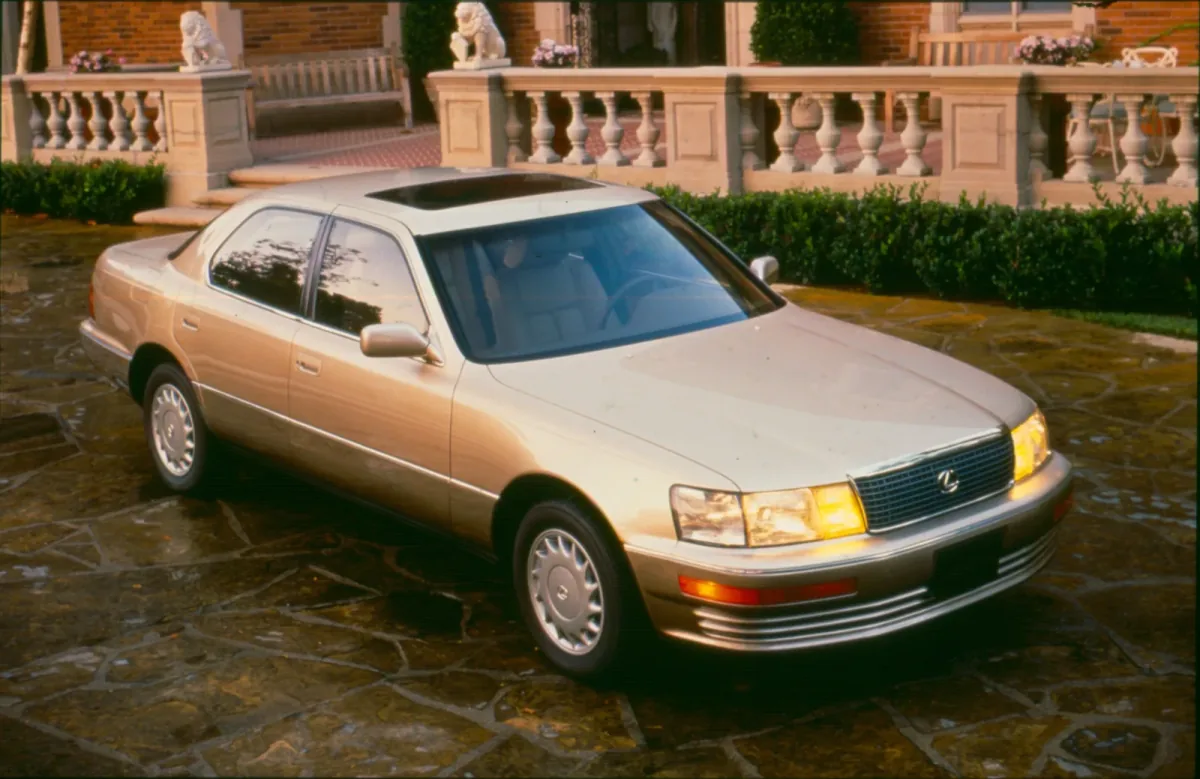 Lexus LS 400 pierwszej generacji stoi na kamiennym podjeździe przed rezydencją — ujęcie z przodu pod kątem, w zachodzącym słońcu, z włączonymi światłami.