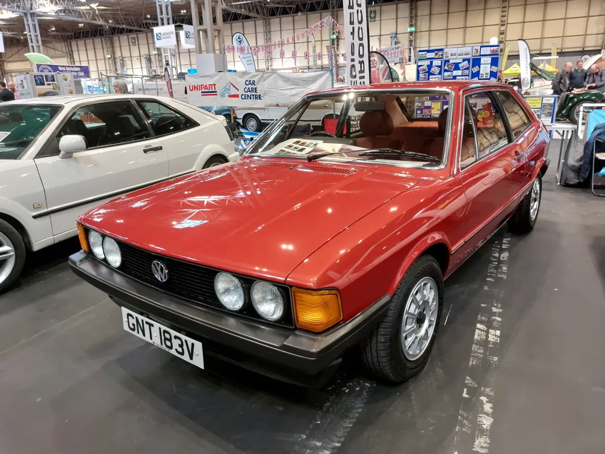 Odrestaurowany Volkswagen Scirocco I 1.6 z 1980 roku w czerwonym lakierze — przód z lewej strony, cztery okrągłe reflektory i czarna atrapa ze znaczkiem VW. Zdjęcie z wystawy NEC Restoration Show w Birmingham, marzec 2022.