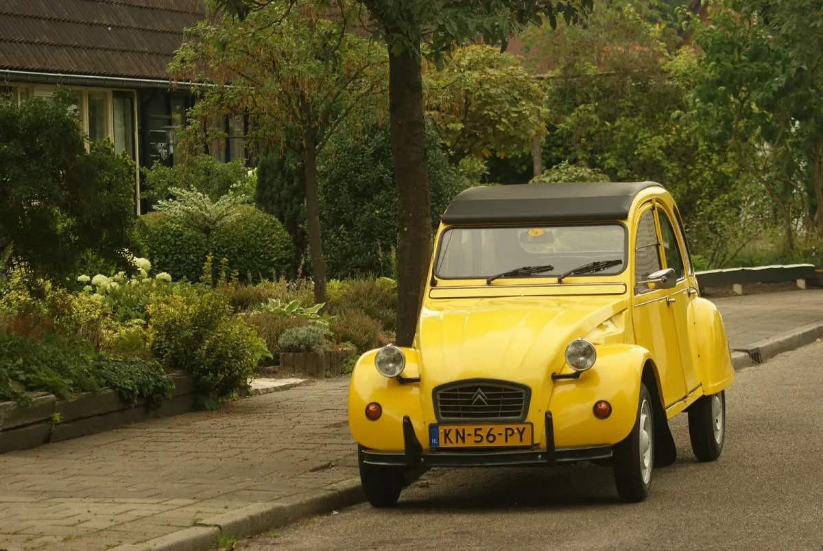 Żółty Citroën 2CV z holenderską żółtą tablicą rejestracyjną (znak NL) zaparkowany przy uliczce wśród zieleni