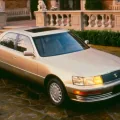 Lexus LS 400 pierwszej generacji stoi na kamiennym podjeździe przed rezydencją — ujęcie z przodu pod kątem, w zachodzącym słońcu, z włączonymi światłami.