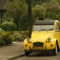 Żółty Citroën 2CV z holenderską żółtą tablicą rejestracyjną (znak NL) zaparkowany przy uliczce wśród zieleni