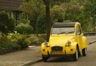 Żółty Citroën 2CV z holenderską żółtą tablicą rejestracyjną (znak NL) zaparkowany przy uliczce wśród zieleni