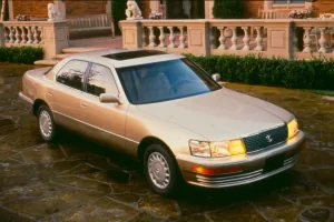 Lexus LS 400 pierwszej generacji stoi na kamiennym podjeździe przed rezydencją — ujęcie z przodu pod kątem, w zachodzącym słońcu, z włączonymi światłami.