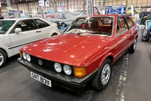 Odrestaurowany Volkswagen Scirocco I 1.6 z 1980 roku w czerwonym lakierze — przód z lewej strony, cztery okrągłe reflektory i czarna atrapa ze znaczkiem VW. Zdjęcie z wystawy NEC Restoration Show w Birmingham, marzec 2022.