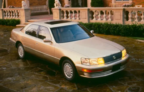 Lexus LS 400 pierwszej generacji stoi na kamiennym podjeździe przed rezydencją — ujęcie z przodu pod kątem, w zachodzącym słońcu, z włączonymi światłami.